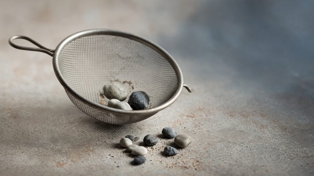 A metal kitchen sieve holding a few stones, with more stones scattered on a grey surface