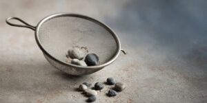 A metal kitchen sieve holding a few stones, with more stones scattered on a grey surface